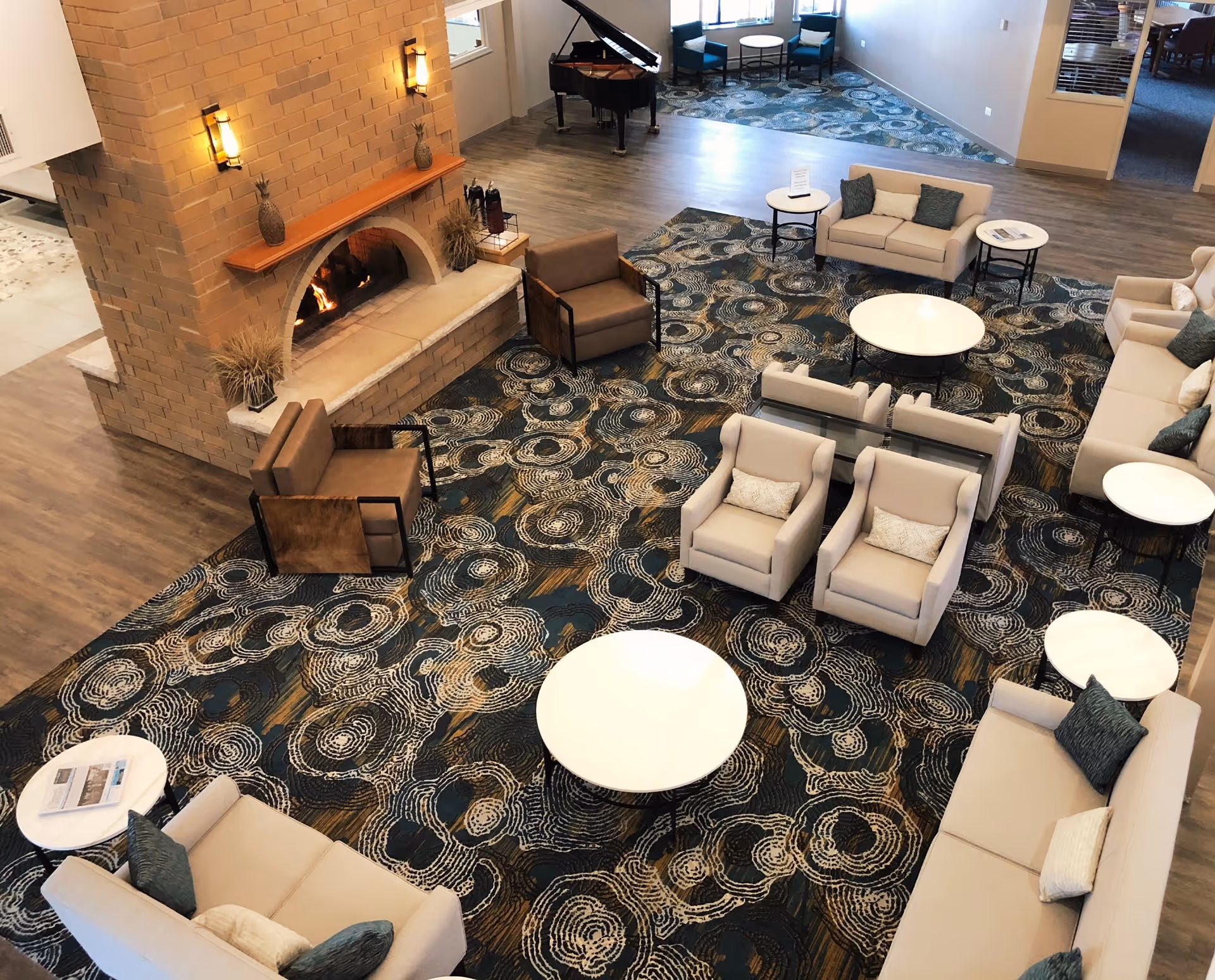 A spacious living room area with multiple beige sofas and armchairs arranged around several round white tables on a patterned carpet. There is a large brick fireplace with a fire burning, flanked by two wall sconces. A grand piano is visible in the background near a window with two blue chairs and a small table.