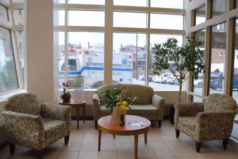 A bright waiting lounge with floral chairs, a couch, and large windows in a senior facility.