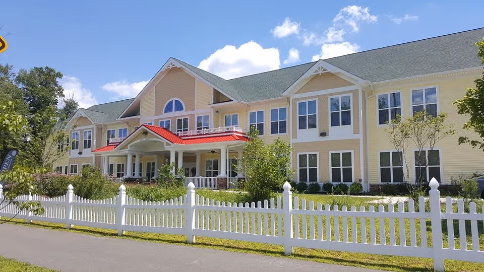 Exterior view of a large two-story yellow building with white trim and a green roof, featuring a covered entrance with a red roof. The building is surrounded by a white picket fence, green shrubs, and trees under a blue sky with some clouds.