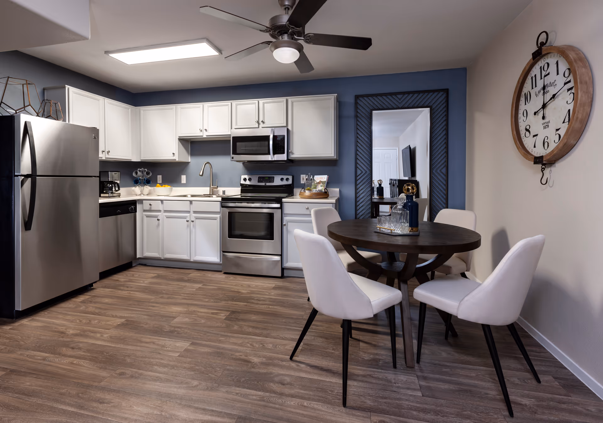 Modern kitchen and dining area with white cabinets, stainless steel appliances including refrigerator, dishwasher, stove, and microwave. A round dark wood dining table with four white upholstered chairs is positioned near a large wall clock and a tall mirror with a dark frame. The floor is wood with a medium brown finish, and the walls are painted in a combination of blue and beige tones.