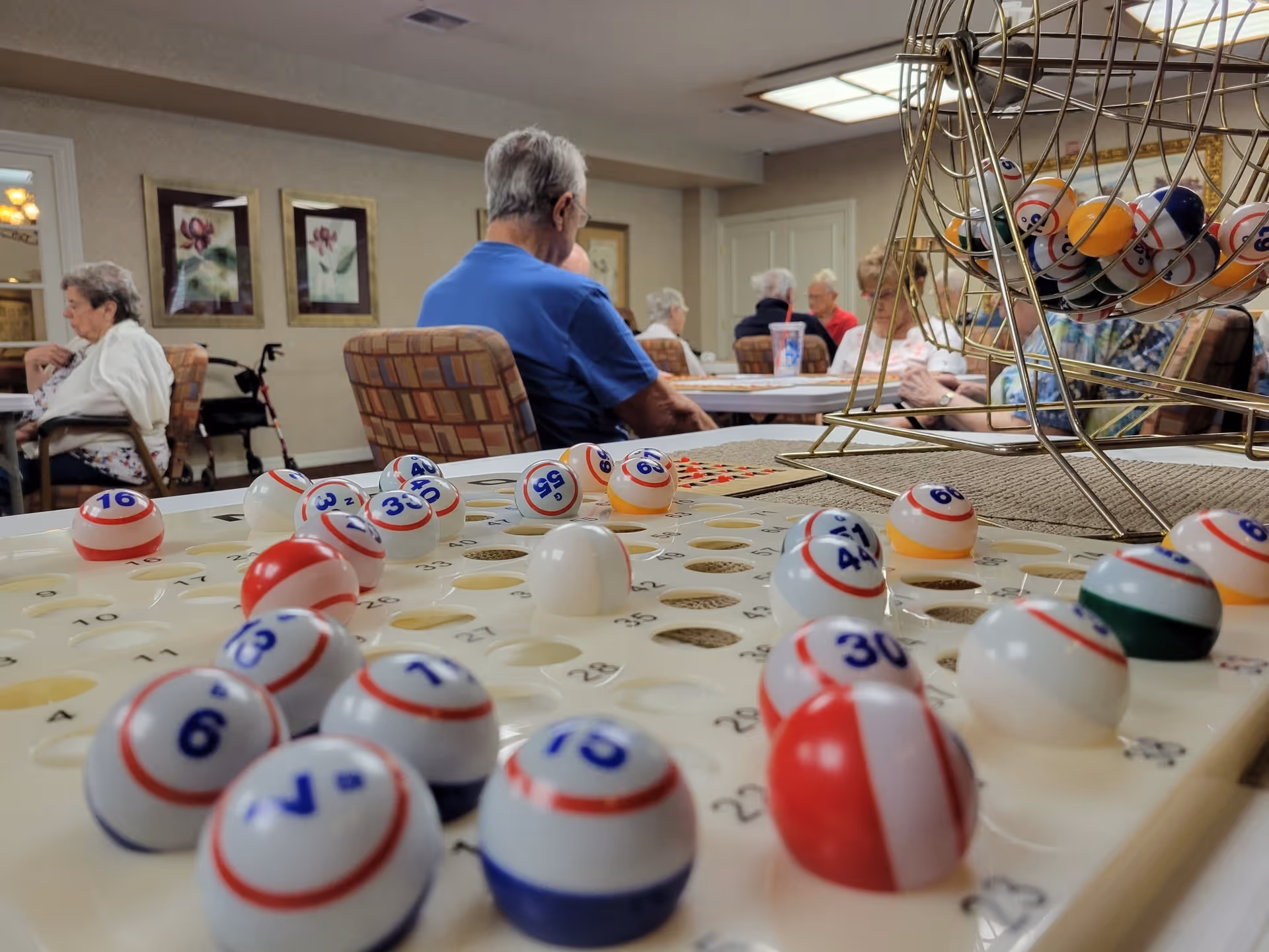 Close-up view of a bingo game with numbered balls on a bingo board and a bingo cage, with elderly people sitting and socializing in the background in a common room.