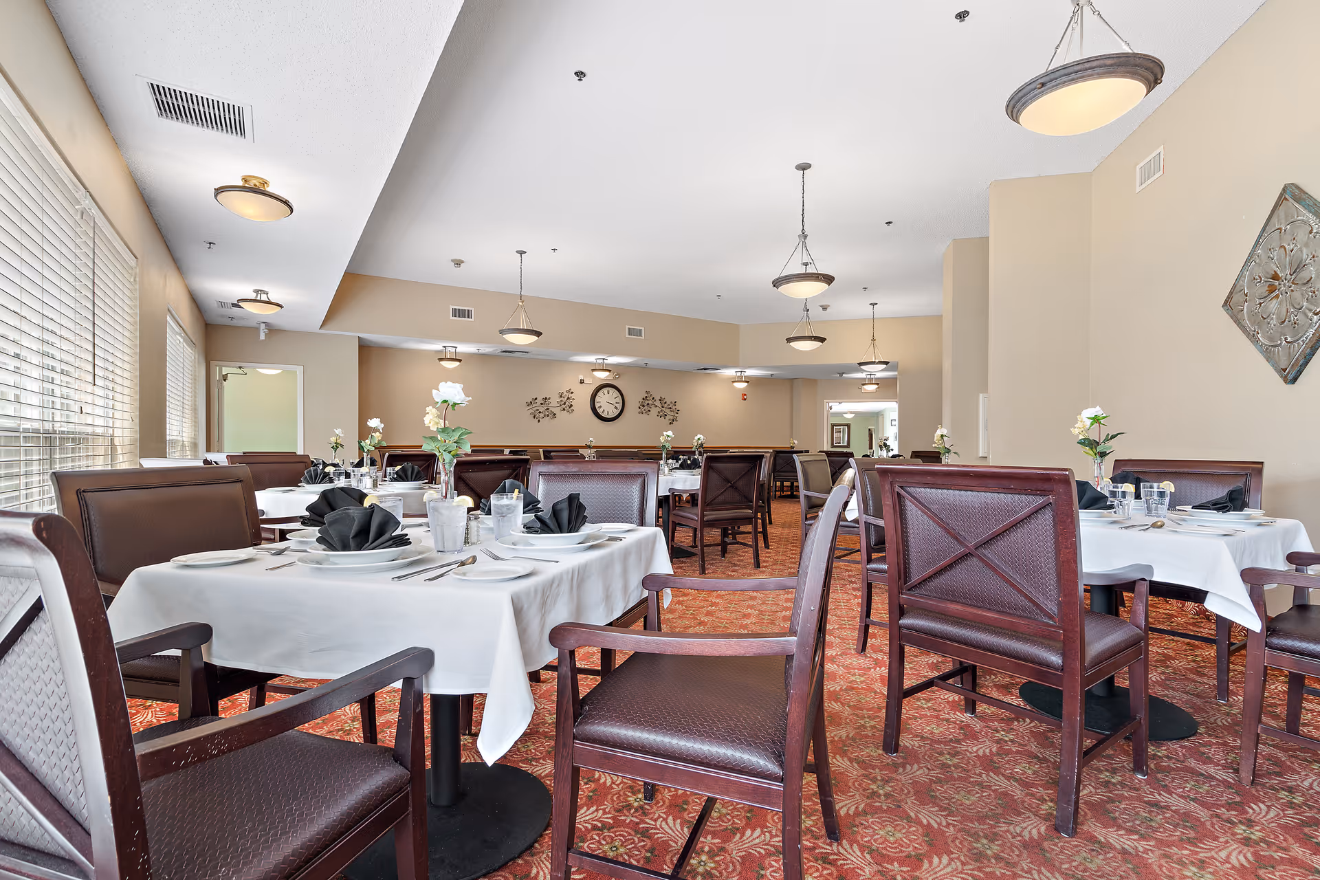 A dining room with multiple tables covered in white tablecloths, each set with plates, glasses, silverware, and black folded napkins. The room has brown chairs with cushioned seats and backs, beige walls, and a patterned red carpet. There are ceiling lights and wall decorations, including a clock and floral wall art.