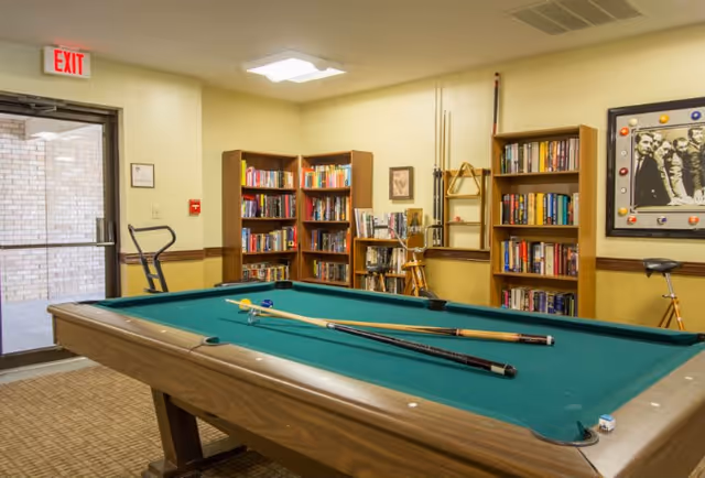 A recreational room with a pool table in the center, two pool cues and a triangle rack on the table. The room has bookshelves filled with books along the walls, a framed black and white picture, and a door with an exit sign above it. The walls are painted beige with a wooden trim.