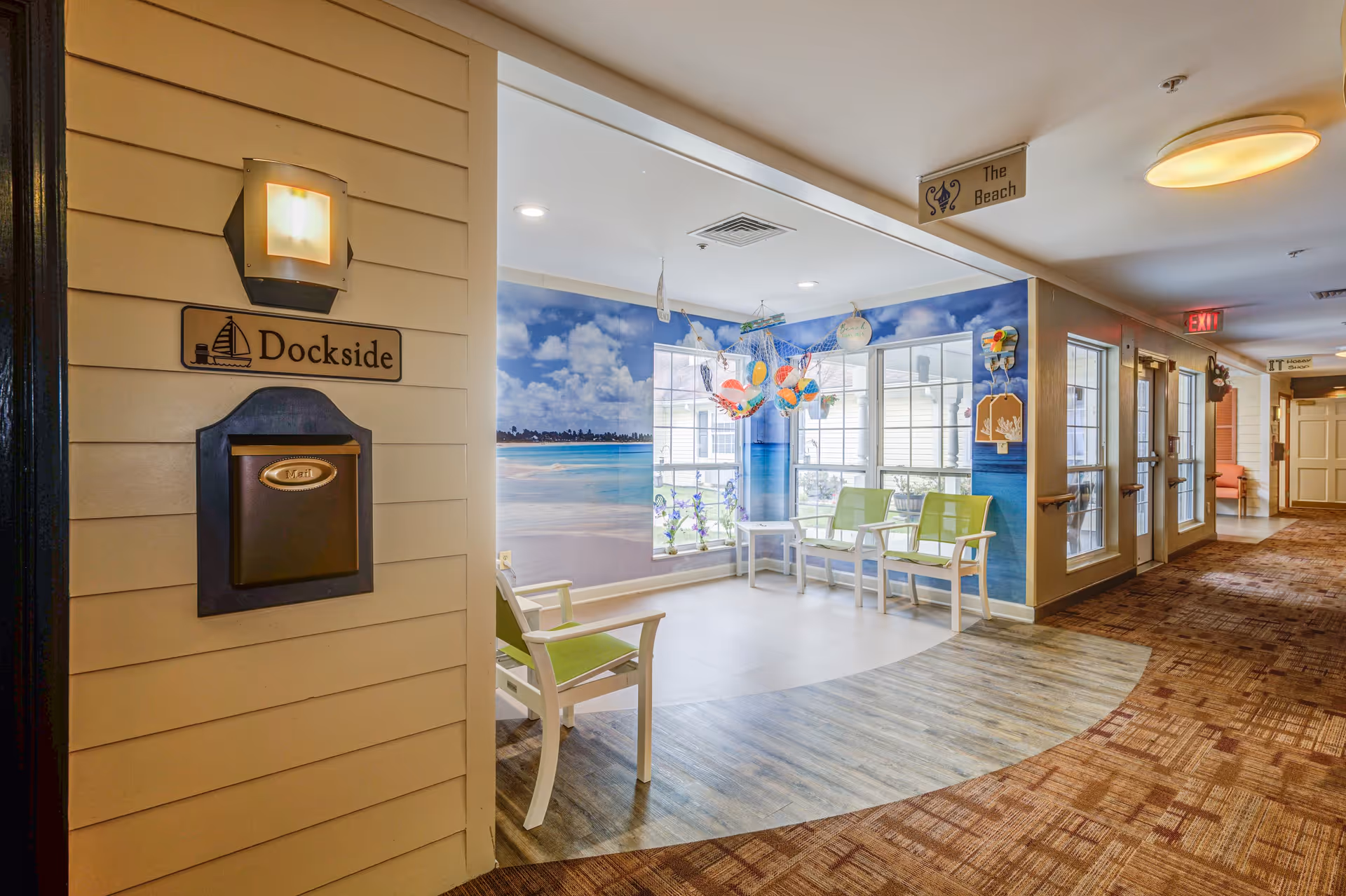 Interior hallway area in a senior living facility with a seating nook decorated with a beach theme. The nook has green chairs, a mural of a beach scene on the wall, and hanging colorful decorations. Signs labeled 'Dockside' and 'The Beach' are visible, along with a mailbox and a wall light fixture. The hallway has carpeted flooring and doors leading to other rooms.
