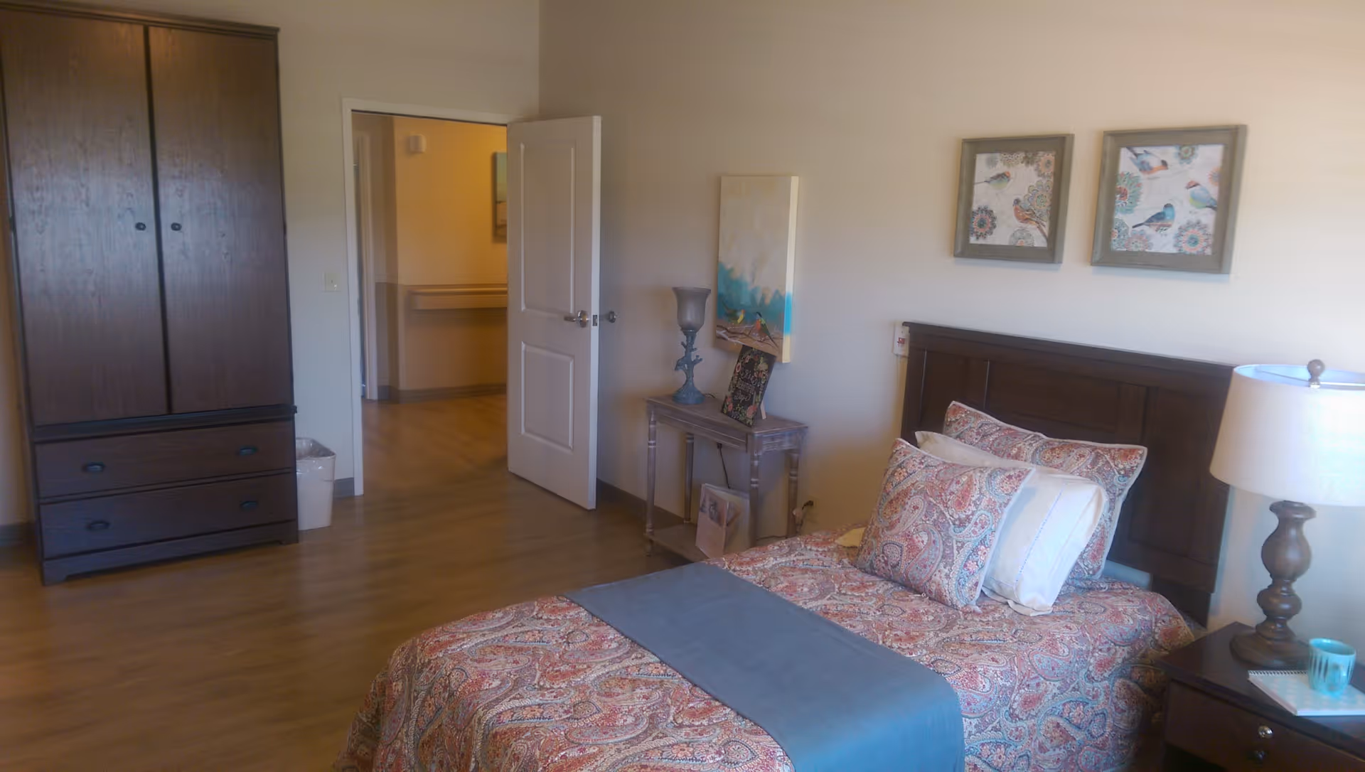 A cozy bedroom with a single bed featuring a patterned bedspread and multiple pillows. There is a wooden nightstand with a lamp and a cup, and a small table with decorative items and artwork on the wall above it. A wooden wardrobe is positioned against the opposite wall, and an open door leads to a hallway with wooden flooring throughout.
