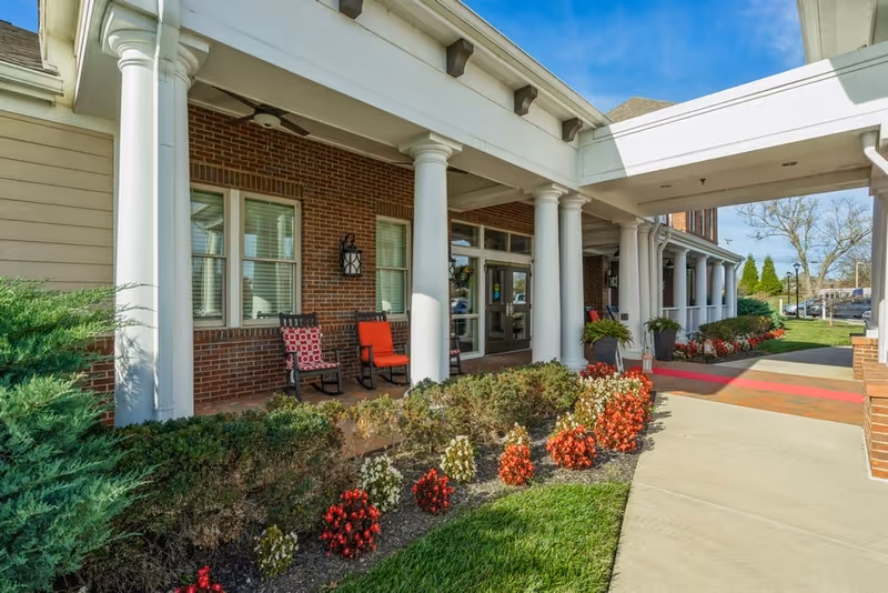 Entrance of a senior living facility with white columns, brick walls, and a covered walkway. There are two chairs with red cushions on the porch, surrounded by neatly trimmed bushes and red and white flowers. The sky is clear and blue.