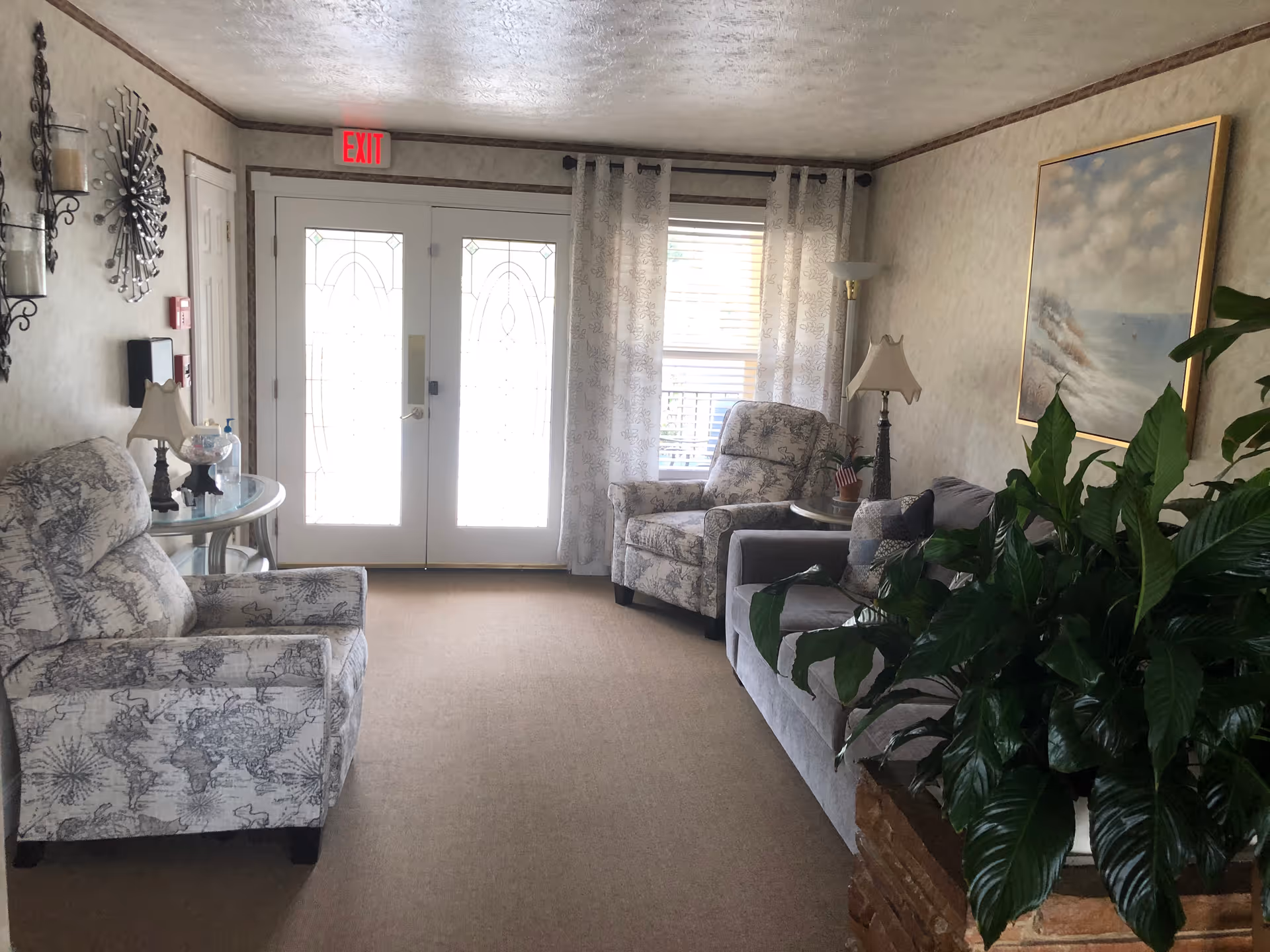 A cozy living room area with two patterned armchairs and a gray sofa with pillows. There is a large green plant in the foreground on the right, a side table with a lamp and small American flag, and a painting of a cloudy sky and ocean on the wall. The room has beige walls, carpeted floor, and double glass doors with an exit sign above them. Sheer curtains cover a window next to the doors.