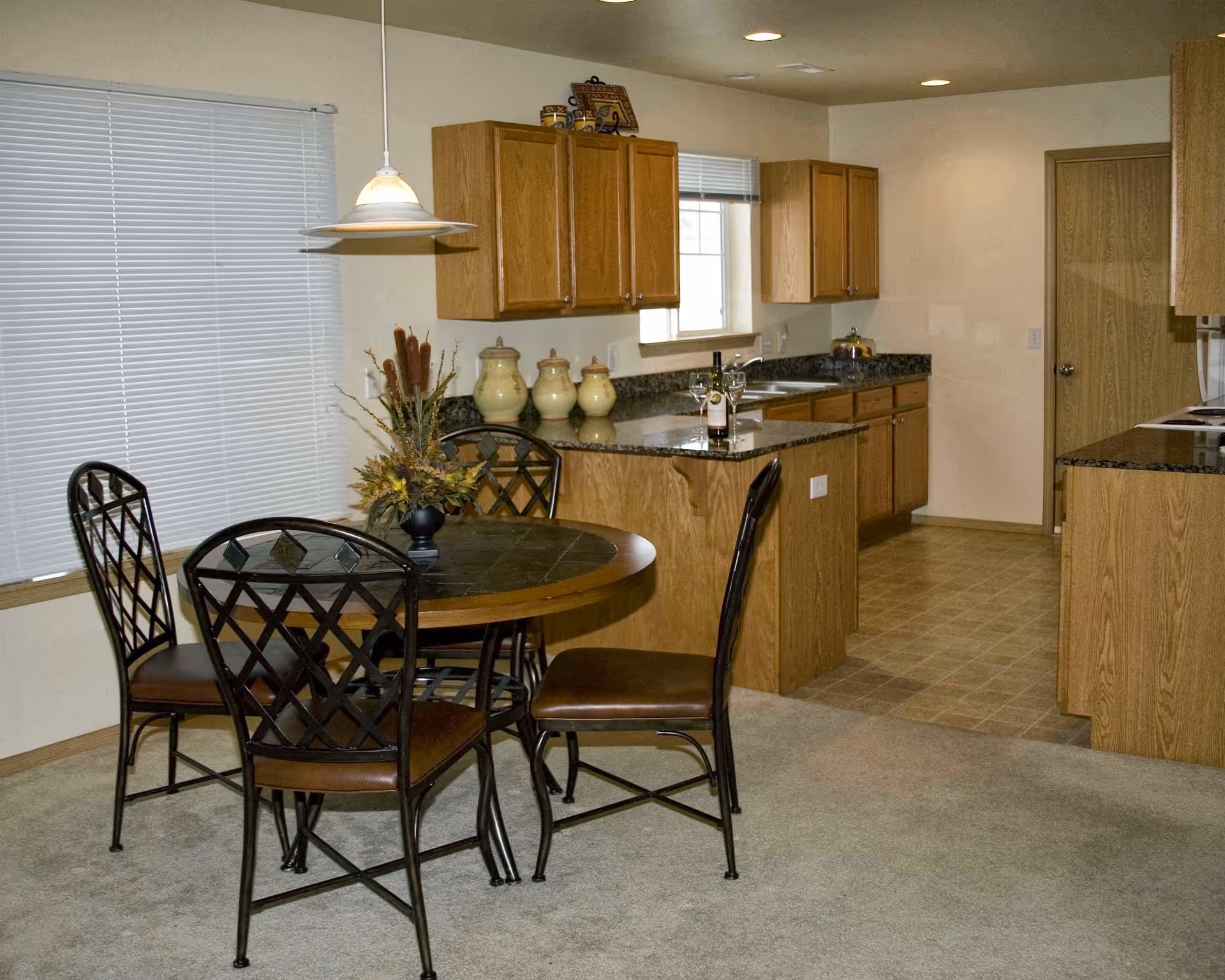 Interior view of a kitchen and dining area in a senior living facility. The dining area features a round table with four metal chairs with brown cushions. A decorative centerpiece is on the table. The kitchen has wooden cabinets, granite countertops, a sink under a window, and various decorative jars on the counter. The floor in the kitchen area is tiled, while the dining area has carpet. A pendant light hangs above the dining table.