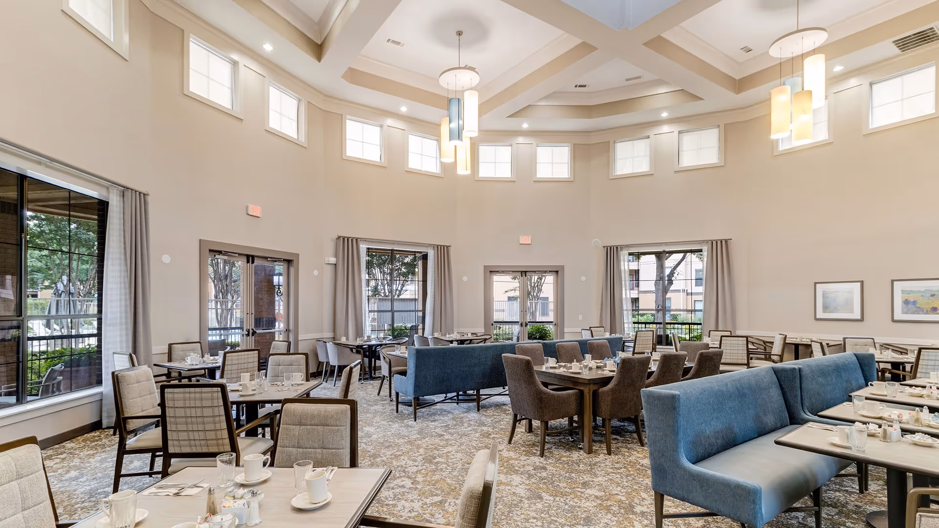 A spacious dining room with multiple tables and chairs arranged neatly. The room features large windows with curtains, allowing natural light to fill the space. There are blue cushioned bench seats and brown upholstered chairs around the tables. The ceiling is high with modern pendant lights hanging down, and the walls are painted in a neutral color with framed artwork.