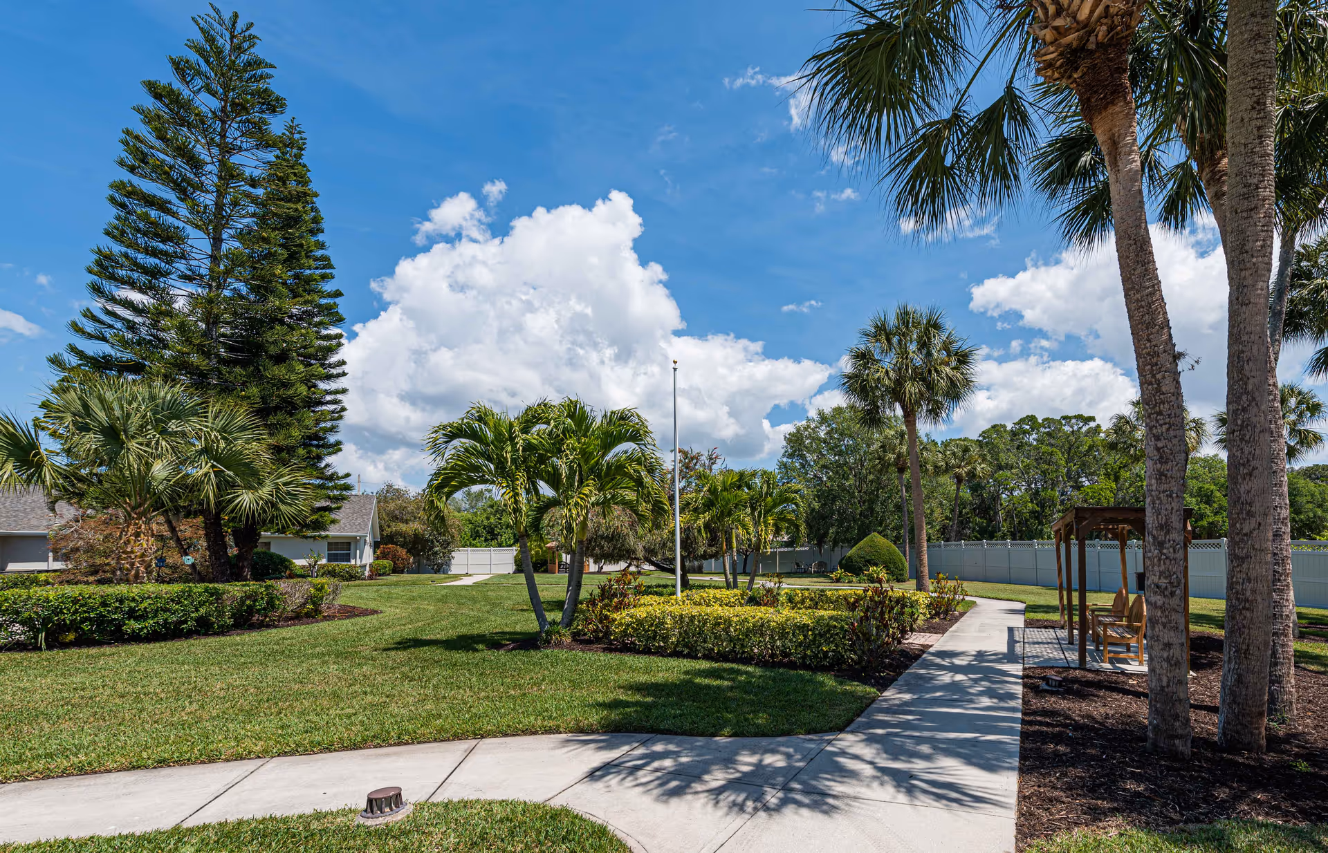 A sunny outdoor garden area at Arden Courts - ProMedica Memory Care Community in Sarasota, featuring a paved walkway surrounded by green grass, palm trees, and other tropical plants. There is a small wooden gazebo with benches on the right side and a bright blue sky with scattered clouds overhead.