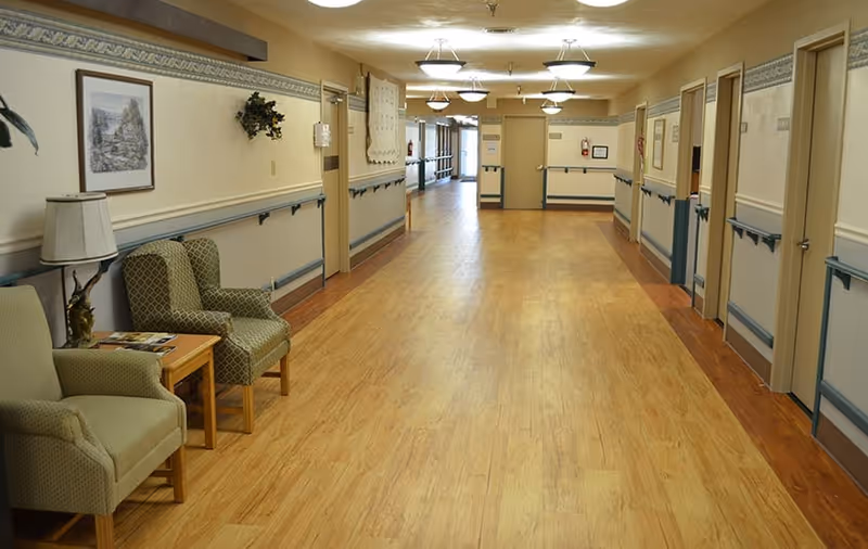 Long, well-lit corridor in a senior living facility with chairs and a small table to the left, handrails and doors lining the hallway.