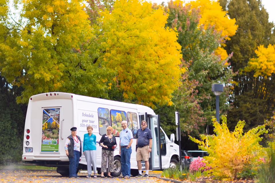 A group of five seniors standing in front of a white shuttle bus with Creekside Village Retirement Residence branding, surrounded by vibrant yellow and green autumn trees in an outdoor setting.