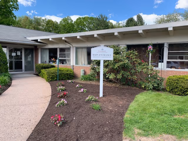 Exterior view of Crestfield Rehabilitation Center & Fenwood Manor showing the main entrance with a pathway leading to the door, surrounded by landscaped bushes, flowers, and green grass under a partly cloudy sky.