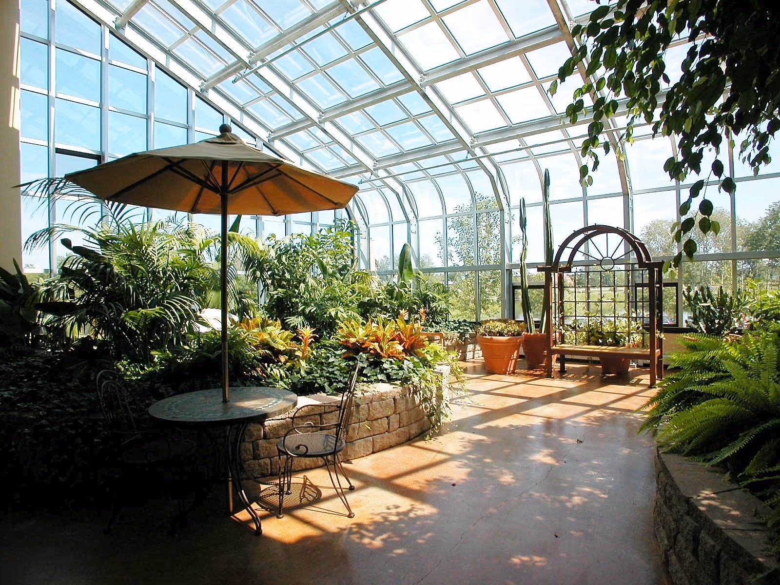 A bright indoor garden room with large glass windows and ceiling allowing sunlight to flood the space. There are various green plants and flowers in raised stone planters. A round metal table with two chairs and a large umbrella is on the left side. On the right, there is a wooden bench with a decorative trellis behind it. The floor is smooth and reflects the sunlight.
