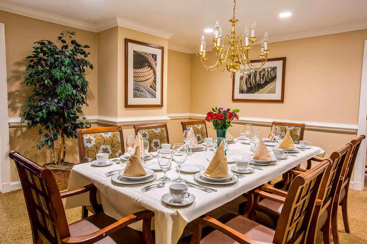 A formal dining room with a rectangular table set for eight people. The table is covered with a white tablecloth and has beige folded napkins, plates, cups, and glasses arranged neatly. A vase with red and purple flowers is placed in the center. The room has beige walls with two framed pictures of ropes and a gold chandelier hanging above the table. There is a large potted plant in the corner.