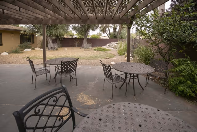 Outdoor patio area with metal tables and chairs under a wooden pergola, overlooking a garden with grass, rocks, and trees.