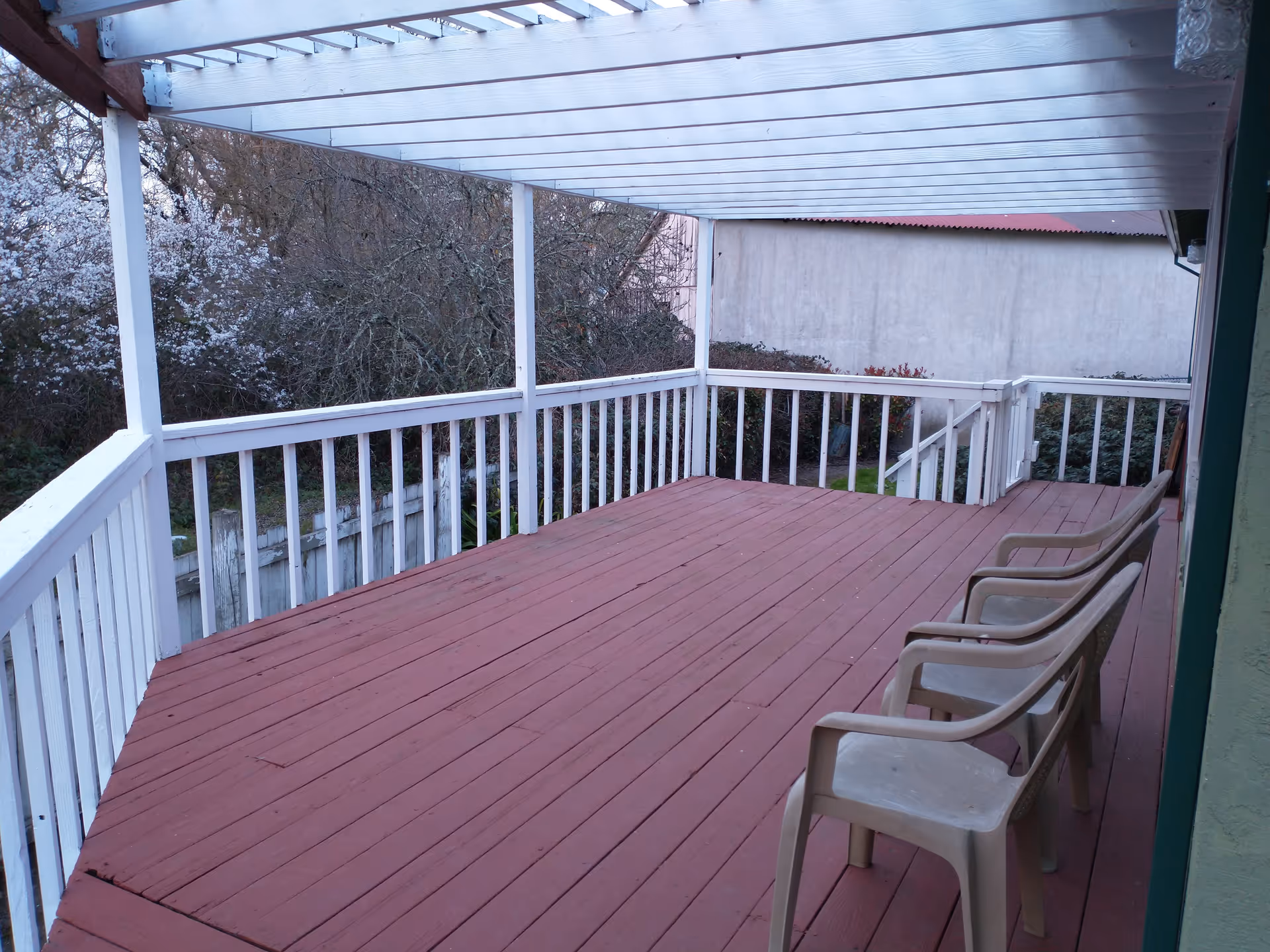 A covered outdoor wooden deck with white railings and a translucent roof. Three plastic chairs are lined up along one side of the deck. Trees and bushes are visible beyond the deck.