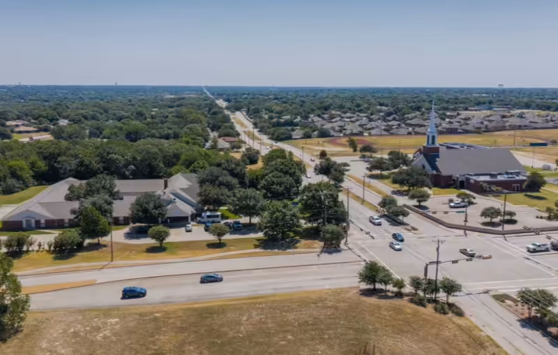 Aerial view of a suburban area showing a senior living facility building surrounded by trees, a nearby church with a tall steeple, roads with cars, and residential neighborhoods extending into the distance under a clear blue sky.