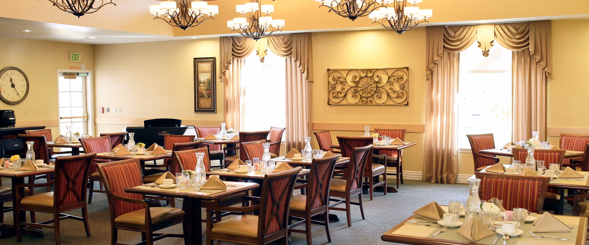 A well-lit dining room with multiple wooden tables and chairs arranged neatly. Each table is set with beige napkins, glassware, cups, and small flower arrangements. The room features large windows with beige curtains, decorative wall art, and chandeliers hanging from the ceiling. A clock and an exit door are visible on one wall.