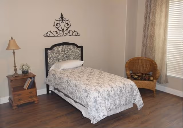 Single-occupancy bedroom with a twin bed covered in floral bedding, a wooden nightstand with a lamp, a wicker chair, and a window with blinds.
