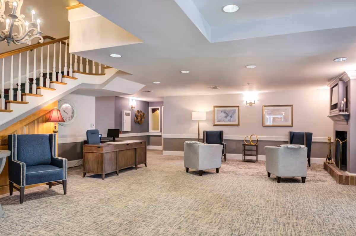 A spacious and well-lit common area in a senior living facility featuring a wooden desk with a computer, several upholstered chairs in blue and gray, a staircase with wooden handrails, a floor lamp, framed artwork on the walls, and a fireplace with a mantle.