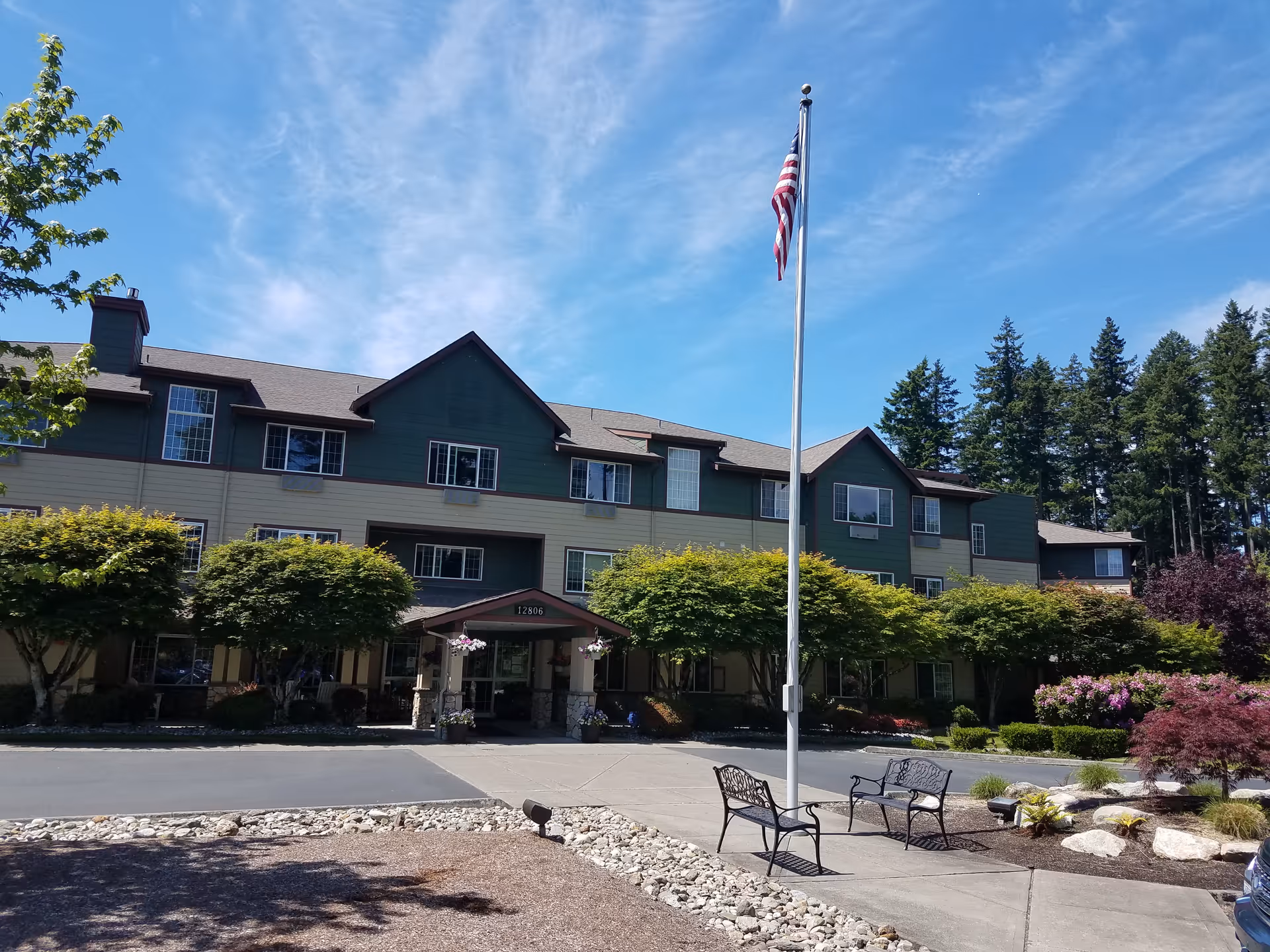 Front exterior of a three-story senior living building with an American flag on a pole, benches, and landscaped trees and shrubs.