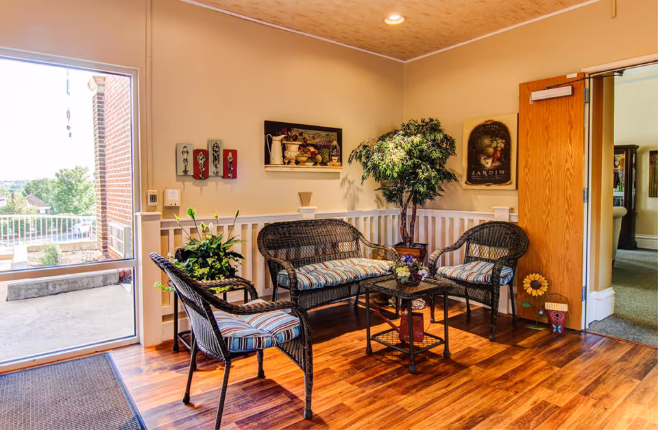 A cozy seating area inside a senior living community featuring a wicker loveseat and two wicker chairs with striped cushions arranged around a small glass-top table. The room has wooden flooring, beige walls adorned with framed artwork, and a potted plant in the corner. A door to the right leads to another room, and a large window on the left shows an outdoor view.
