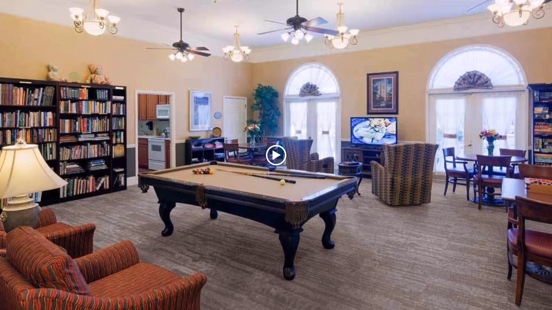 A spacious common room with a pool table in the center, surrounded by comfortable seating including armchairs and dining tables. There is a large bookshelf filled with books on the left side, a television on a stand near two large arched windows with sheer curtains, and a small kitchen area visible through an open doorway. The room is well-lit with ceiling fans and hanging light fixtures.