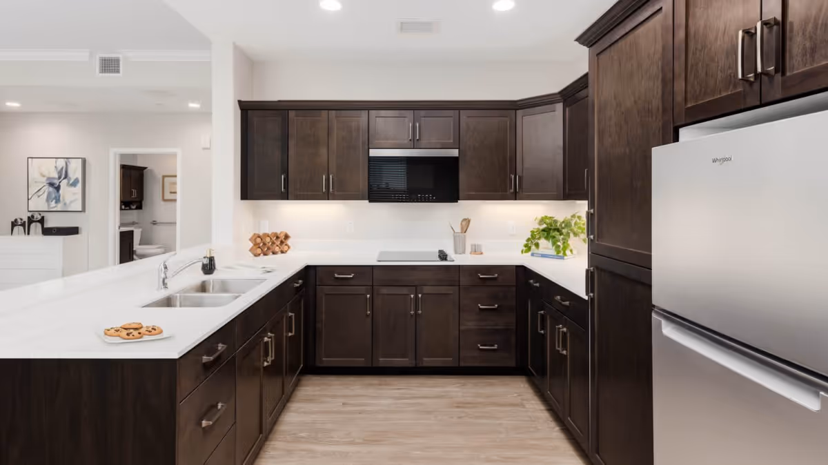 Modern kitchen with dark wood cabinets, white countertops, a stainless steel Whirlpool refrigerator, a built-in microwave, and a double sink. There is a plate of cookies on the counter, a small wine rack, and a green plant on the countertop. The kitchen floor is light wood, and the adjacent room with a toilet is visible in the background.