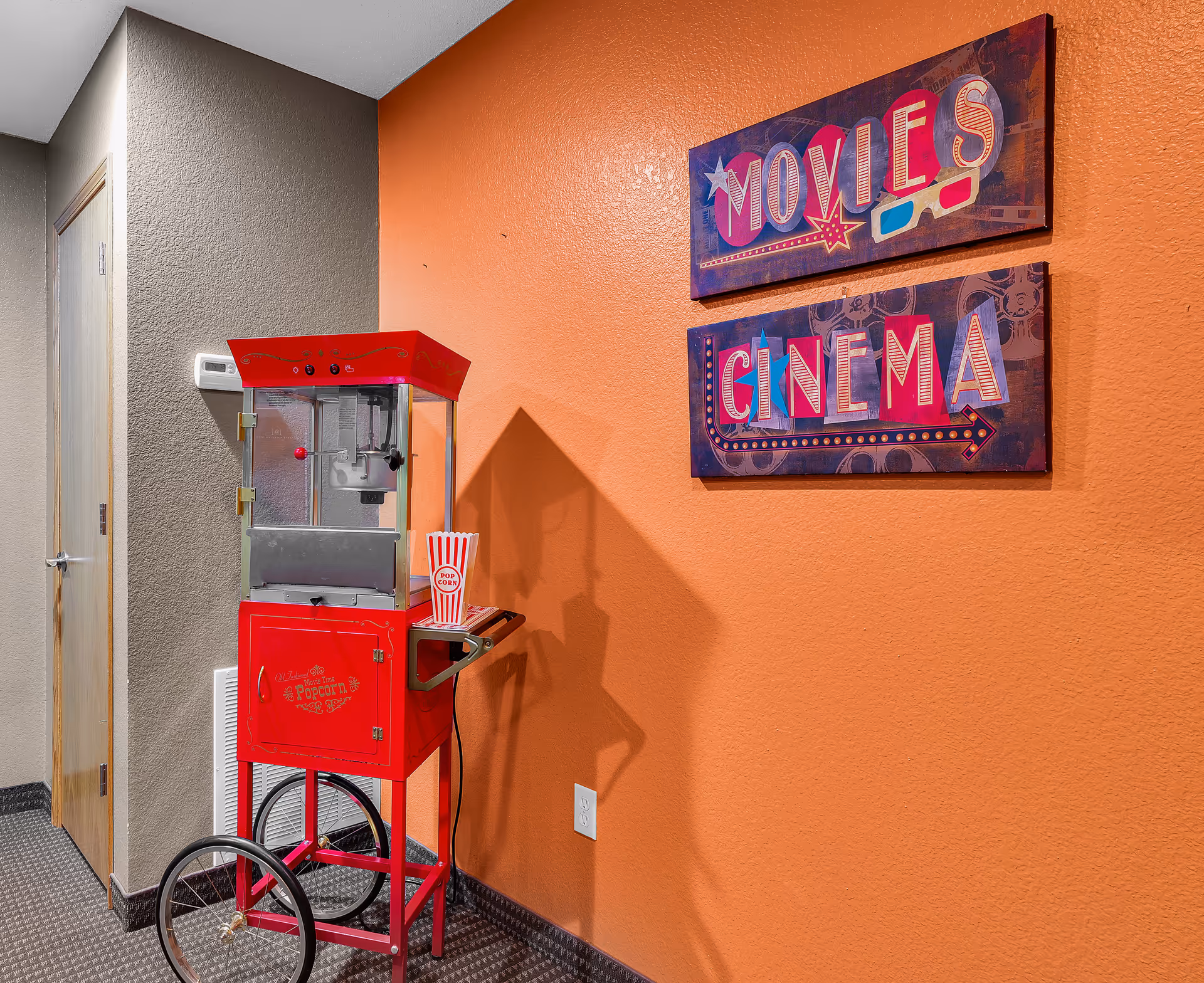 A red vintage-style popcorn machine on wheels is placed against an orange wall. Above it, two colorful signs read 'MOVIES' and 'CINEMA'. The adjacent wall is painted gray with a wooden door and a thermostat visible.