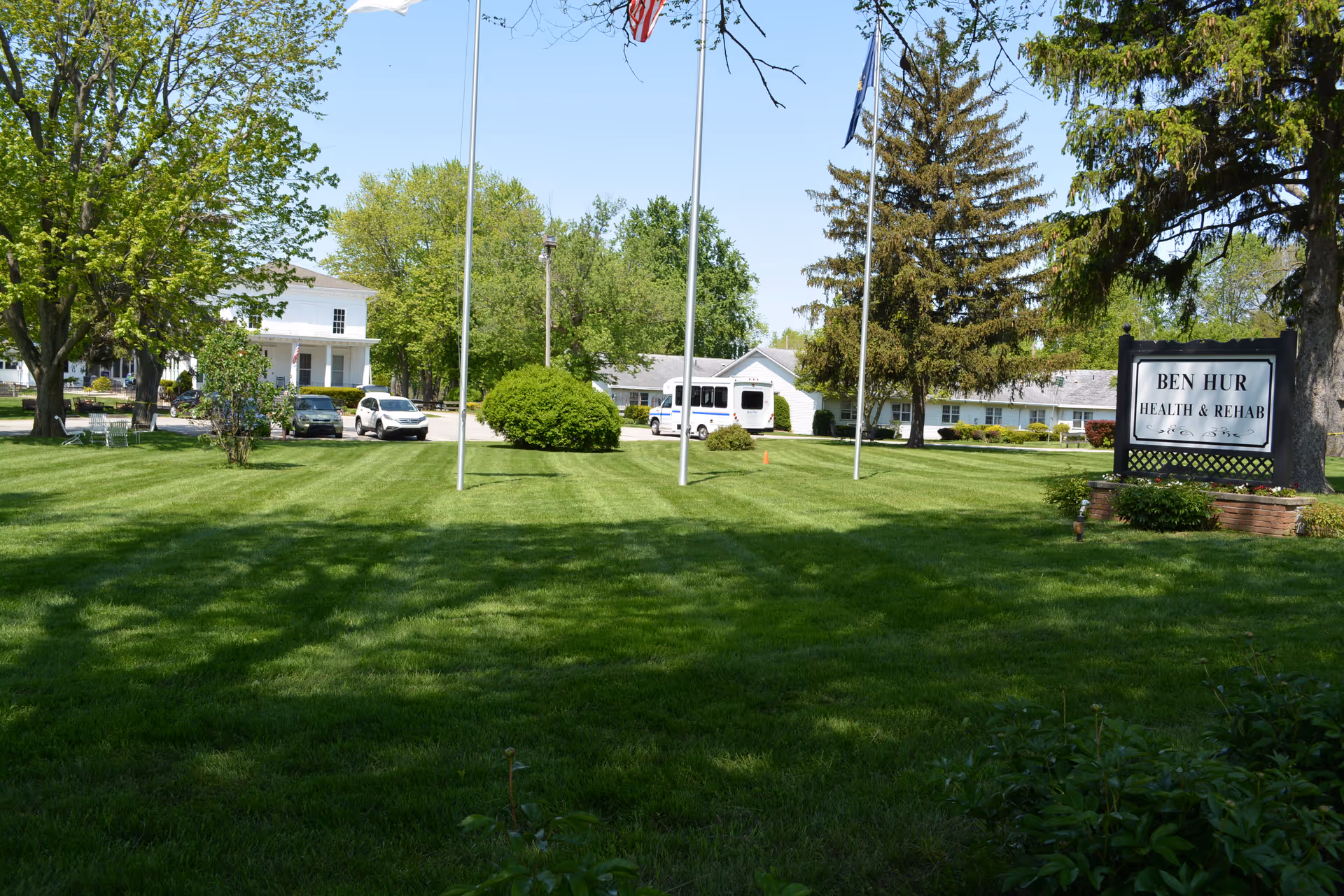 Front lawn with flagpoles and a sign reading "Ben Hur Health & Rehab" in front of the facility buildings.