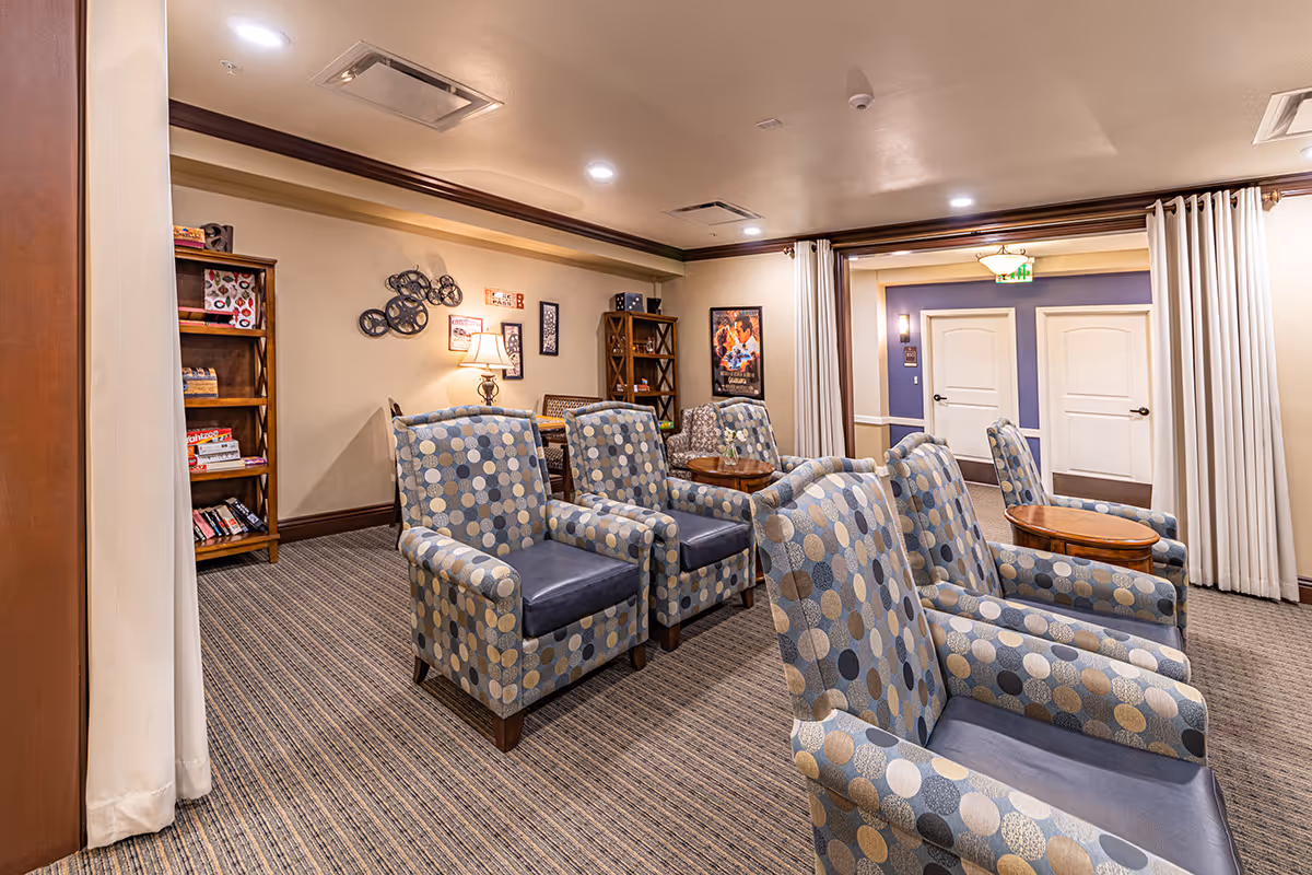 A cozy senior living common area with several patterned armchairs arranged in rows facing a small wooden table. The room has beige walls, carpeted floor, and wooden bookshelves filled with board games and books. There are framed pictures and decorative wall art, with soft lighting from ceiling lights and a table lamp.