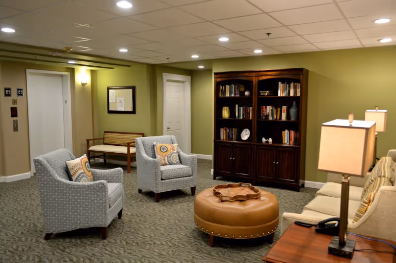 A cozy living room area with two patterned armchairs, a round leather ottoman, a beige sofa with striped pillows, a wooden bookshelf filled with books and decorative items, a wooden bench near an elevator, and two table lamps providing warm lighting.
