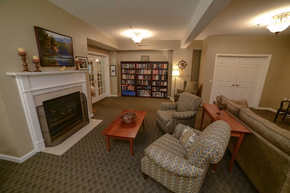 A cozy senior living community lounge featuring a fireplace with candles and a painting above it, two patterned armchairs, a wooden coffee table with a decorative basket, a sofa, a bookshelf filled with books, a floor lamp, and neutral-toned walls and carpet.