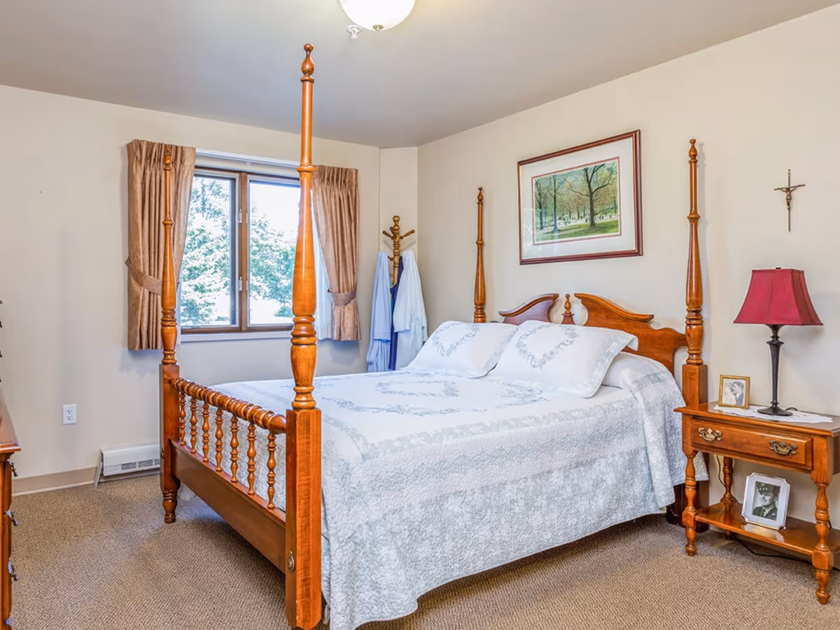 A bedroom with a wooden four-poster bed covered with a white and light blue patterned quilt and matching pillows. There is a window with brown curtains, a wooden nightstand with a red lampshade, framed photos, and a crucifix on the wall. A framed picture of trees hangs above the bed.