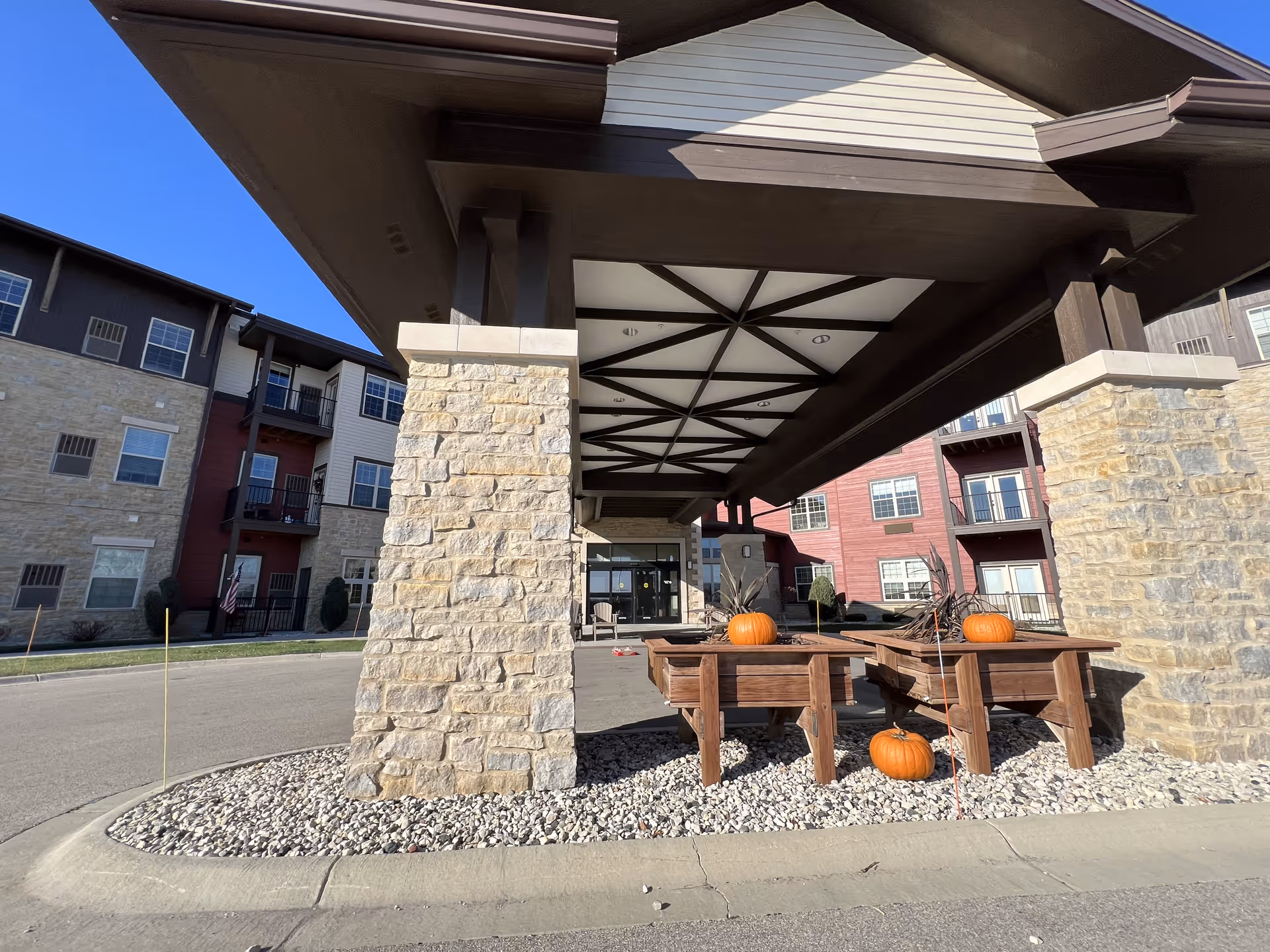 Entrance area of a senior living facility with a covered drop-off zone supported by stone pillars. There are wooden planter boxes with pumpkins placed on and around them, and multi-story residential buildings with balconies in the background under a clear blue sky.