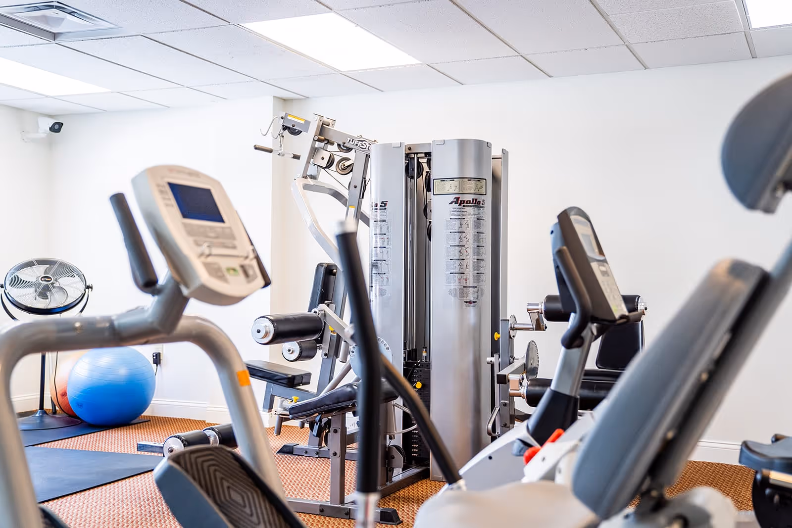 A fitness room with various exercise equipment including stationary bikes, a multi-function weight machine, a blue exercise ball, and a standing fan. The room has white walls and a ceiling with recessed lighting.