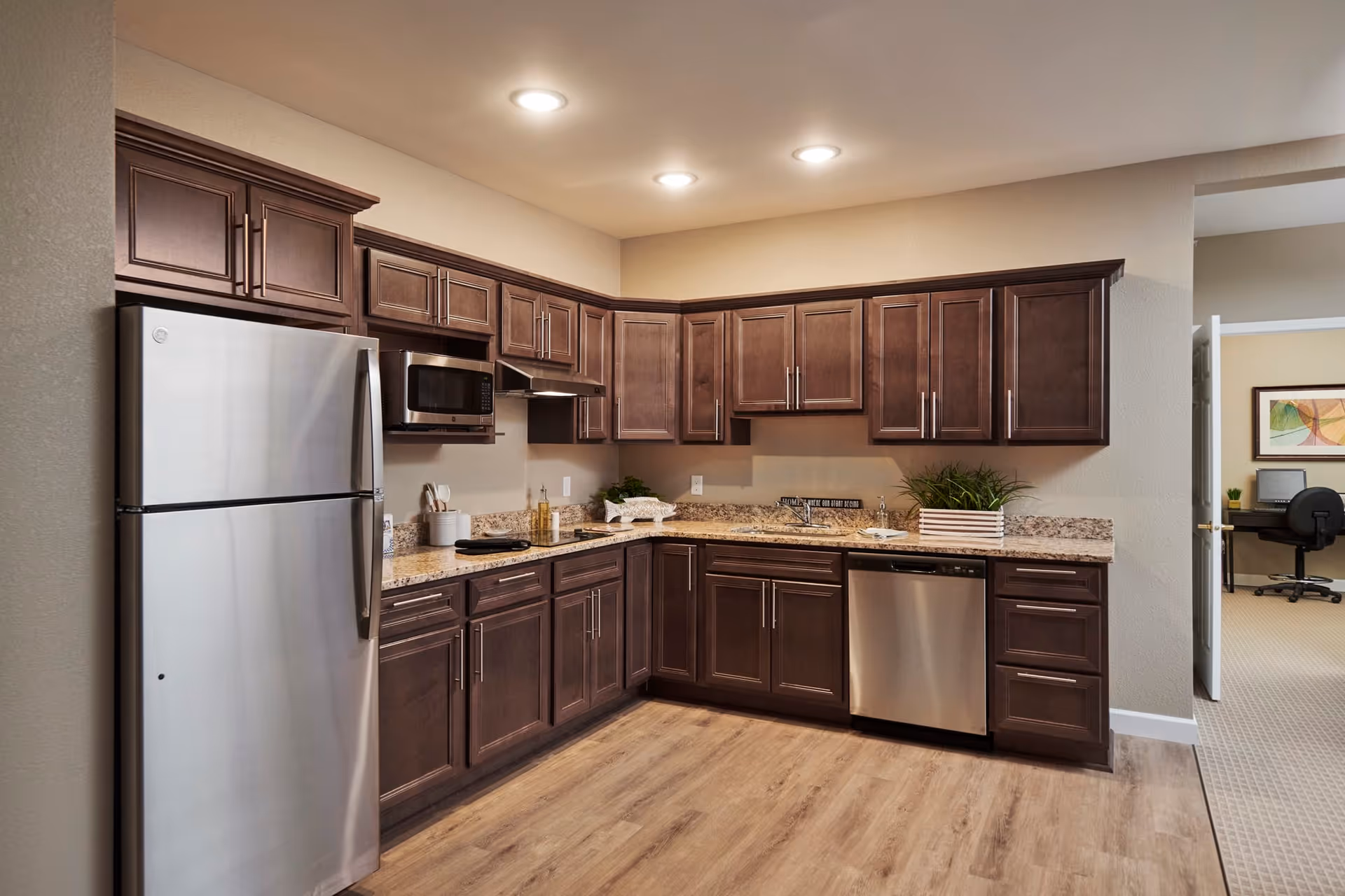 Modern kitchen with dark wood cabinets, granite countertops, stainless steel refrigerator, microwave, dishwasher, and a sink. The kitchen has recessed lighting and wood flooring. An adjacent room with a desk and chair is visible through an open doorway.