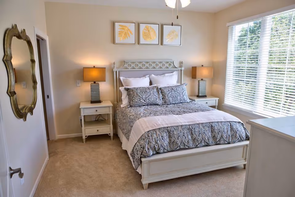 A cozy bedroom with a white wooden bed frame and a patterned bedspread. There are two matching nightstands with lamps on either side of the bed. Above the bed, three framed pictures of golden leaves are hung on the wall. A large window with white blinds lets in natural light, and a decorative mirror is mounted on the wall near the door.