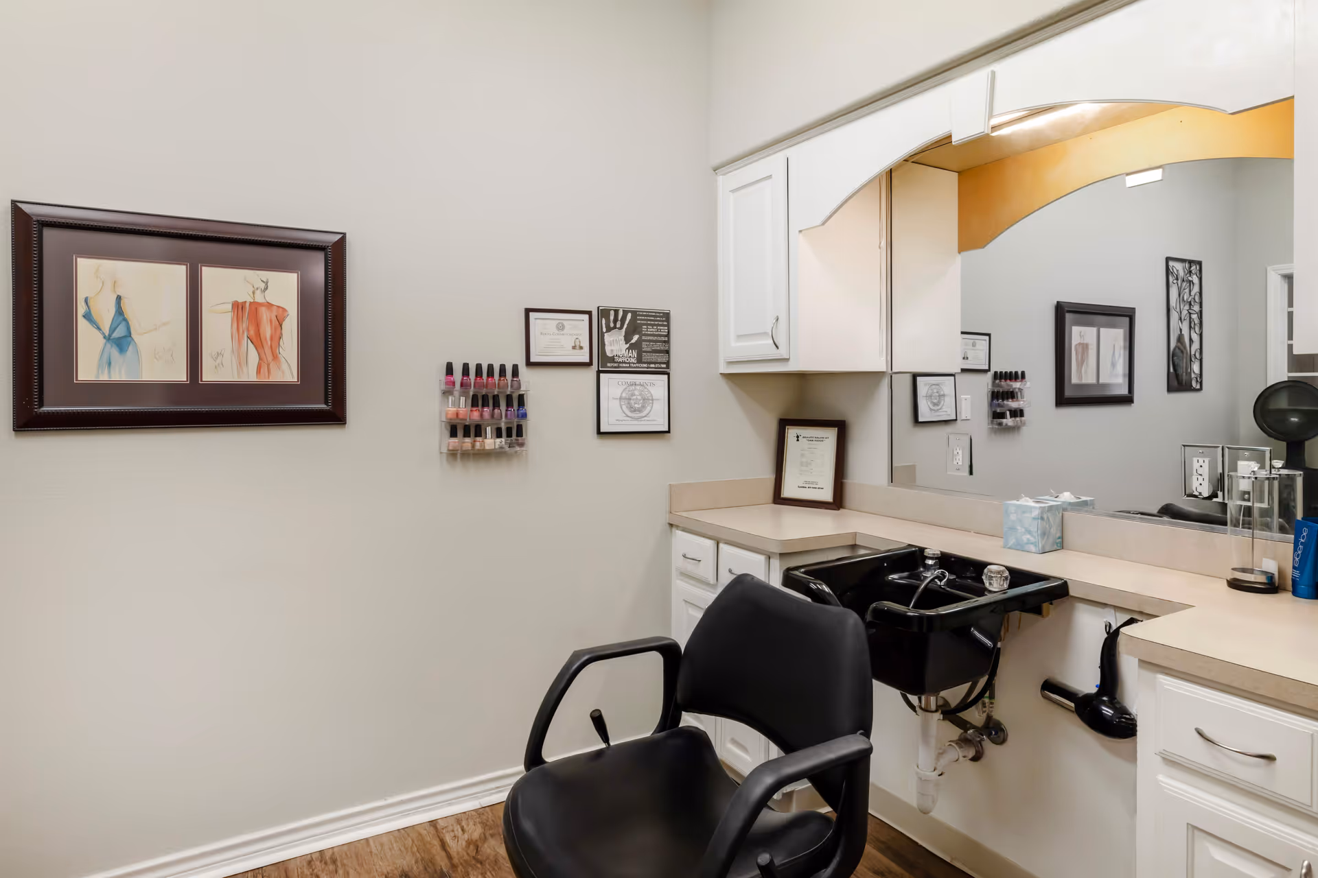 Interior view of a salon area in a senior living facility with a black salon chair in front of a black sink. The countertop has a tissue box and hair care products. There are white cabinets above and below the counter, a large mirror, and framed artwork and certificates on the wall. A rack with nail polish bottles is mounted on the wall.