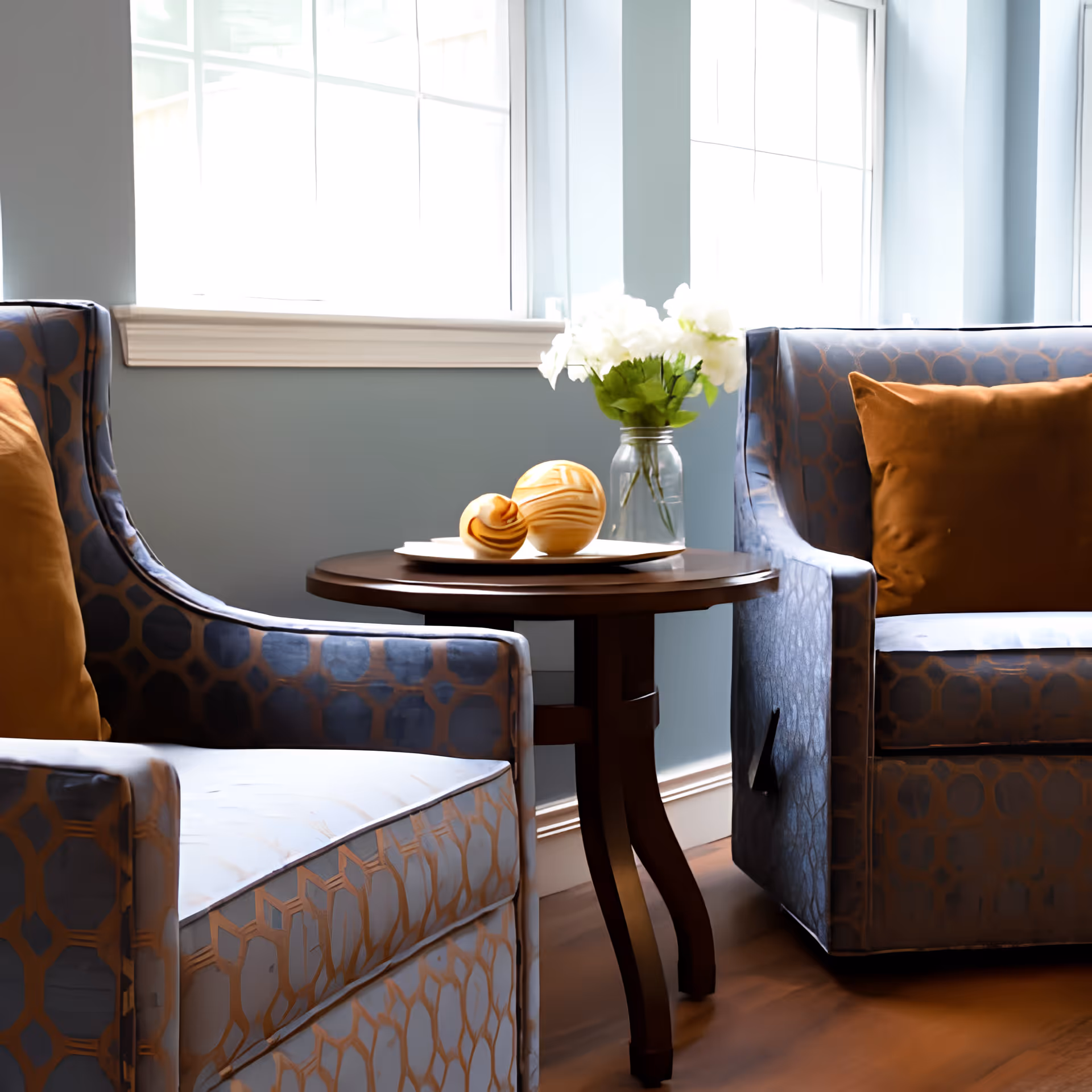 Two patterned armchairs flank a small round wooden side table topped with decorative spheres and a vase of white flowers in front of bright windows.