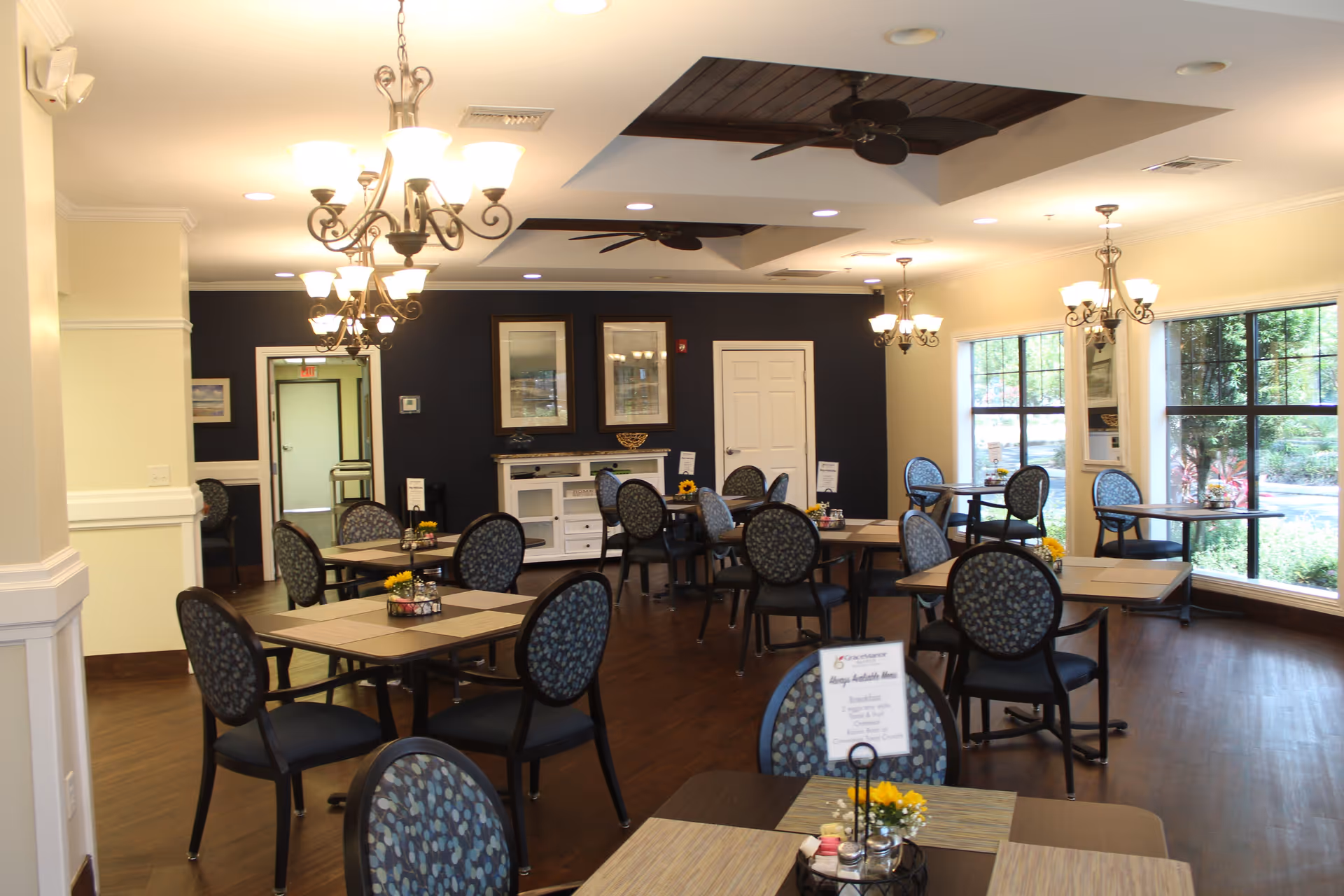A bright and spacious dining room with multiple tables and chairs arranged neatly. The room features large windows letting in natural light, elegant chandeliers hanging from the ceiling, and dark wood flooring. The walls are painted in light and dark tones, and there are framed pictures on the far wall. Small flower arrangements and placemats decorate the tables.