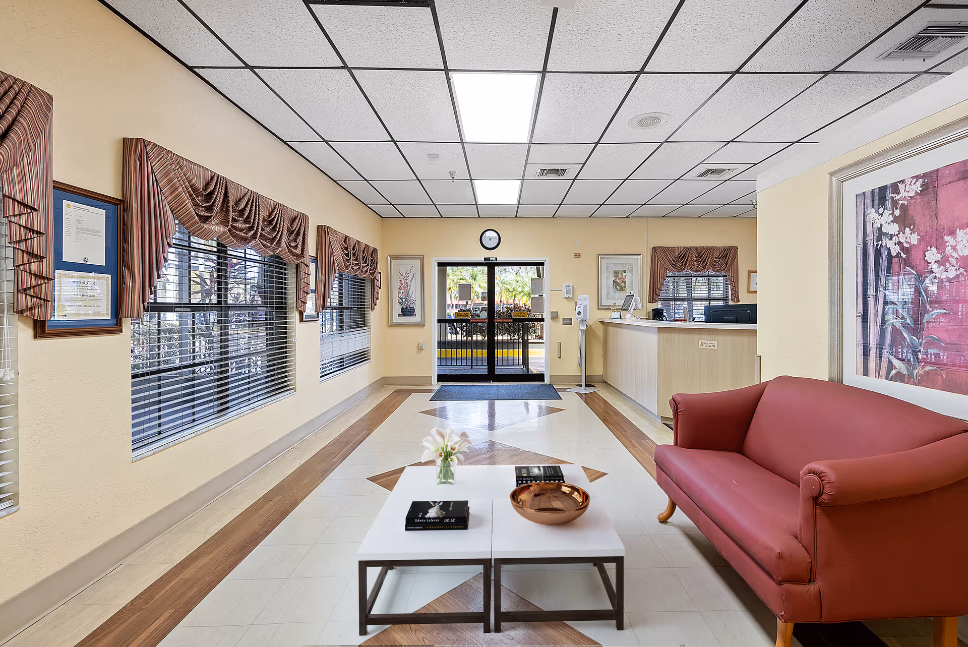 A bright and clean reception area with a red couch on the right, a white coffee table with books and a decorative bowl in the center, and a reception desk in the background. The room has large windows with striped valances, framed artwork on the walls, and a clock above the entrance door.