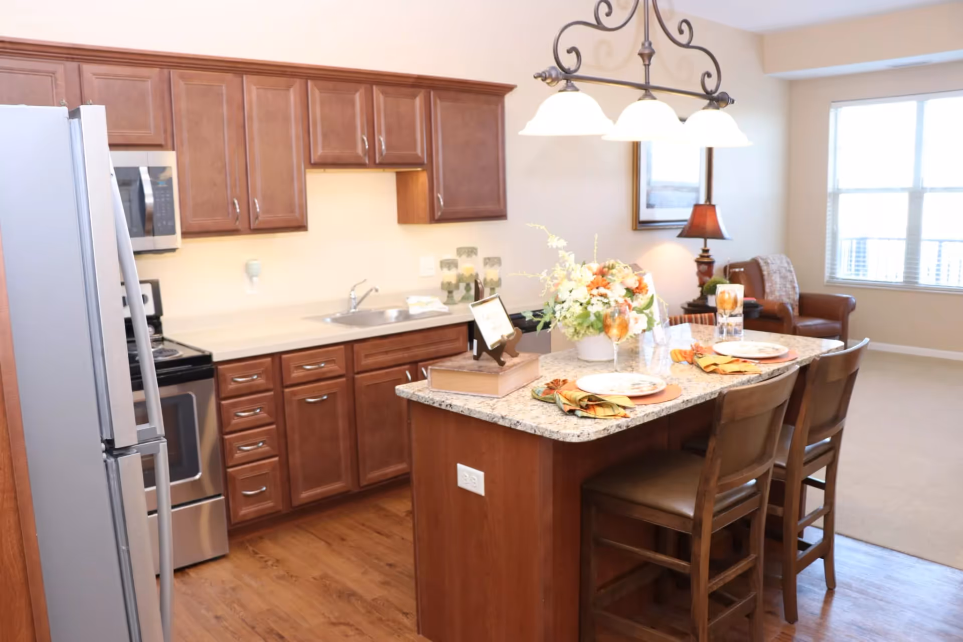 A bright kitchen area with wooden cabinets, a stainless steel refrigerator and oven, and a granite countertop island set with two place settings, napkins, and glasses. A floral centerpiece and a small framed sign are on the island. In the background, there is a living area with a leather chair, a lamp, and a large window letting in natural light.
