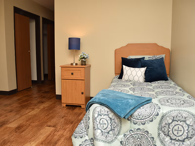 A small bedroom with a single bed featuring a patterned bedspread and several pillows. Next to the bed is a wooden nightstand with a blue lamp and a small vase of flowers. The room has wooden flooring and beige walls, with an open doorway leading to another area.