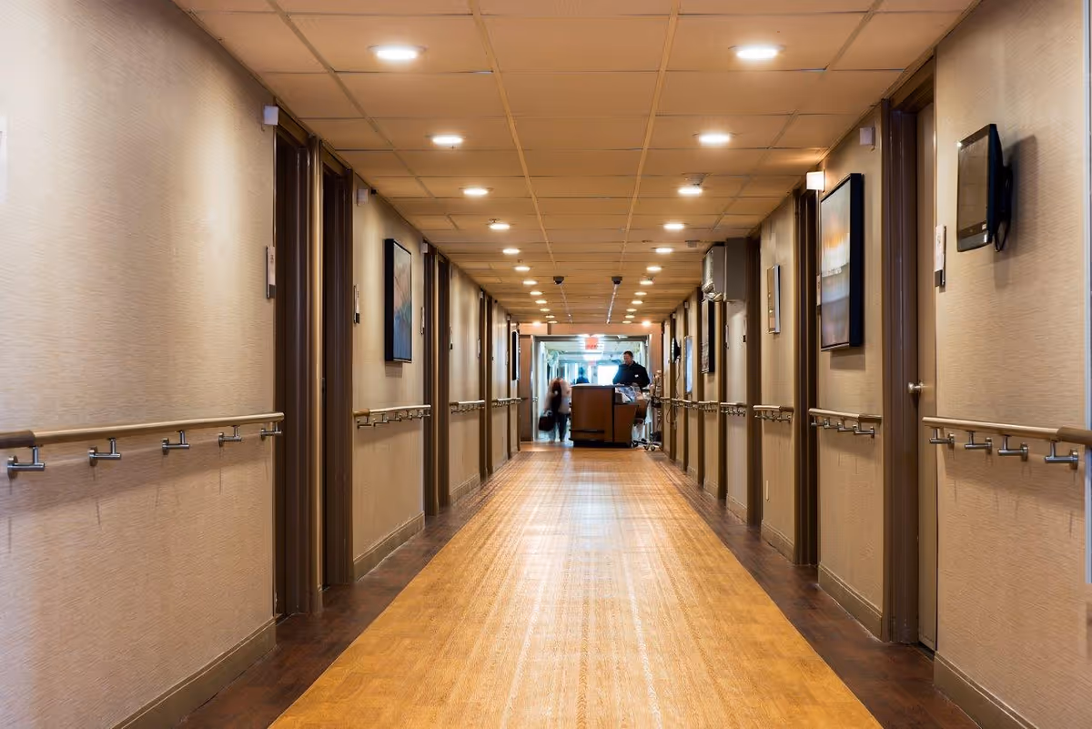 Long hallway in a senior living facility with wooden floors, beige walls, handrails on both sides, multiple doors, framed artwork on the walls, and ceiling lights. Two people are visible at the far end of the hallway near a cart.