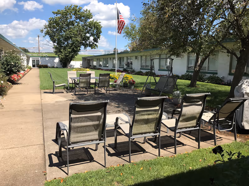 Outdoor patio area with several metal mesh chairs arranged in rows and around tables. There is a concrete walkway, green grass, flower beds, and trees providing shade. An American flag is flying on a flagpole in the center of the patio. The building with white walls and green roof surrounds the patio area under a partly cloudy sky.
