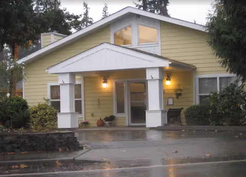 Front exterior view of a single-story yellow building with a white covered entrance supported by columns, two lit wall lanterns on either side of the entrance door, surrounded by greenery and trees on a wet pavement.