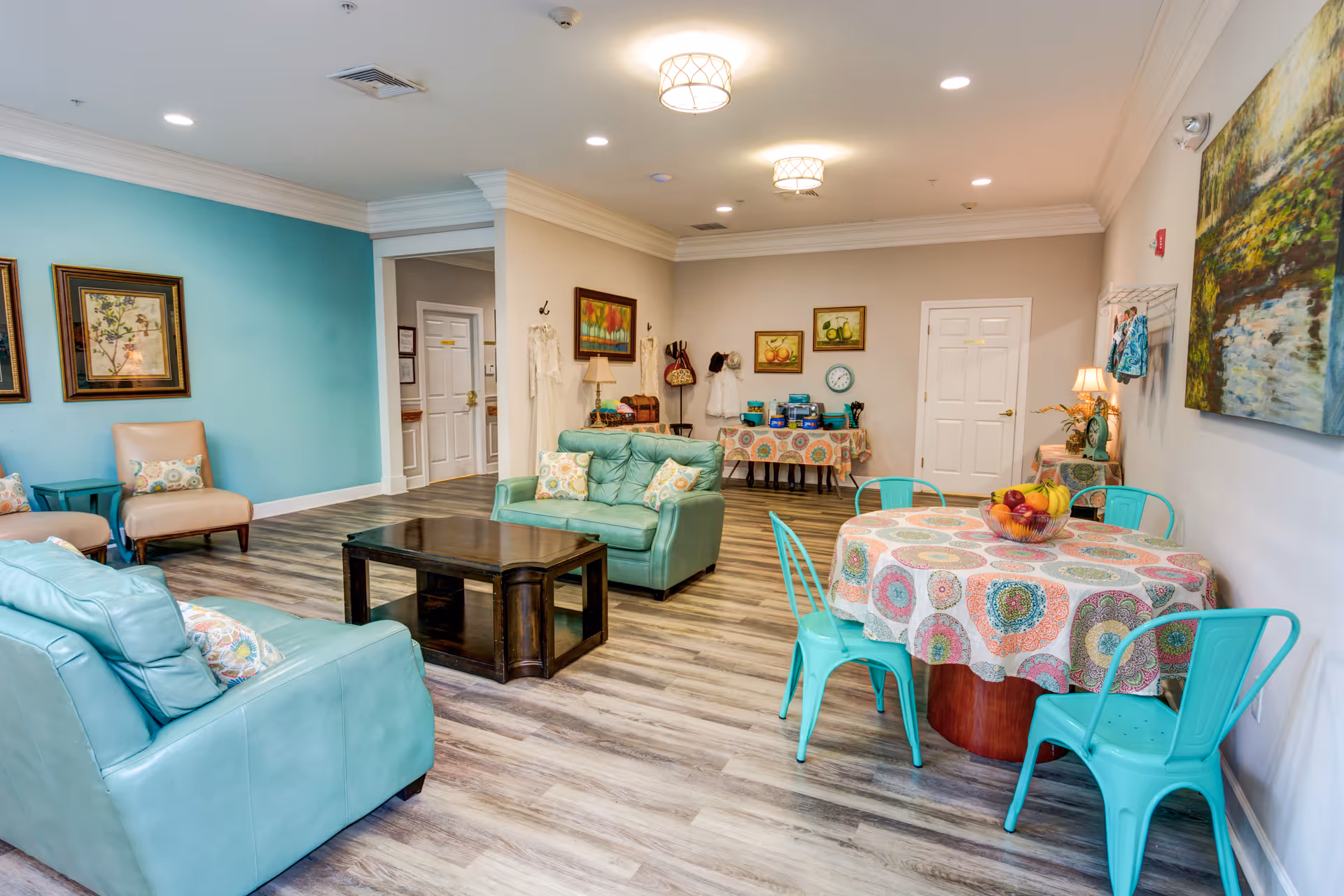 Bright communal living room with teal sofas, a round table covered by a patterned tablecloth surrounded by turquoise chairs, a wooden coffee table, and artwork on the walls.