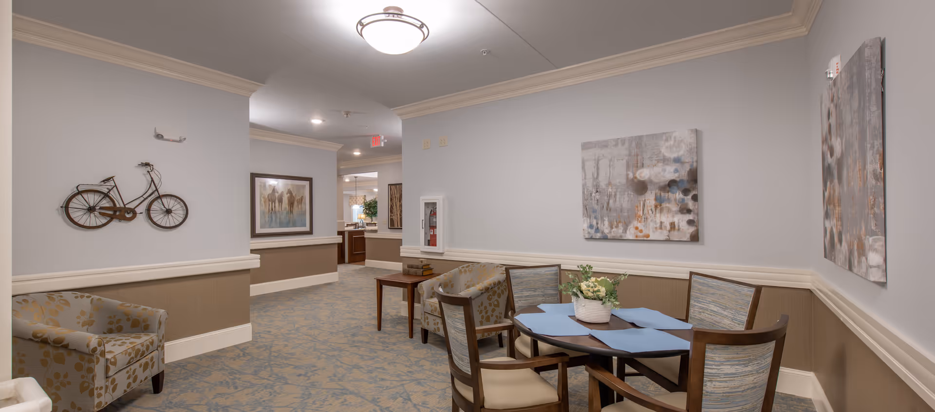 A cozy interior common area with a round table and four chairs, two upholstered armchairs, a decorative bicycle wall art, and abstract paintings on the walls. The space has soft lighting, beige and light blue walls, and patterned carpet flooring.